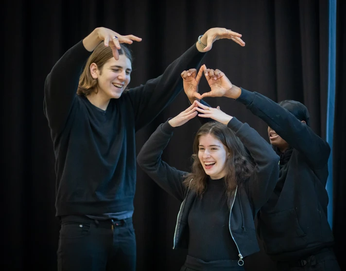 Three students building a creative pose with their hands and arms in a collaborative, playful performance moment during class. Three students building a creative pose with their hands and arms in a collaborative, playful performance moment during class.